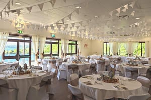 A bright room with bunting on the ceiling, lake views from the windows and circular tables with white table cloths set up for meal.