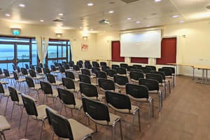 Cream and black chairs set in rows facing a projector screen. There are large windows with lake views.